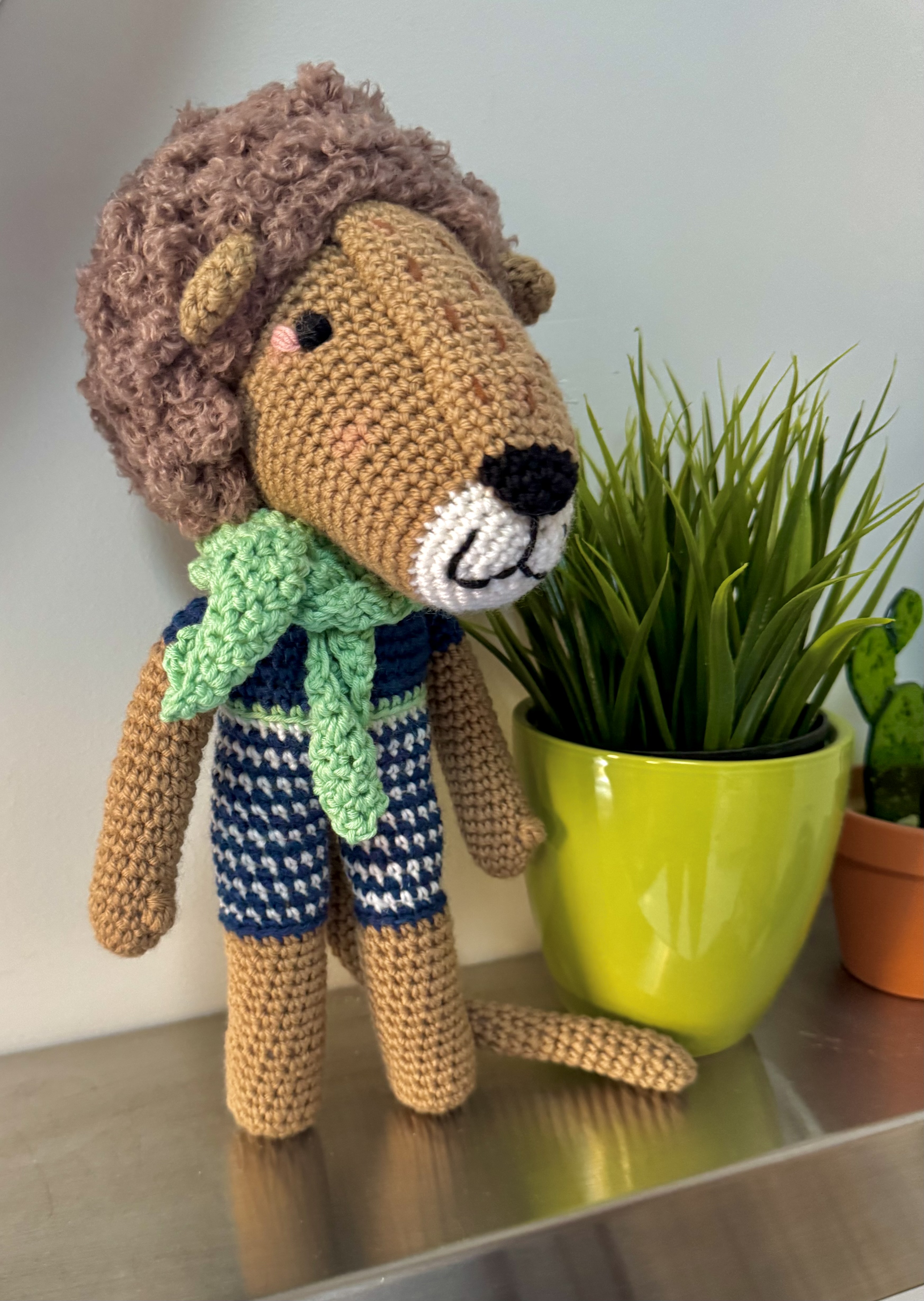 Crocheted lion plush wearing a green scarf, standing on a metallic surface next to two potted plants.