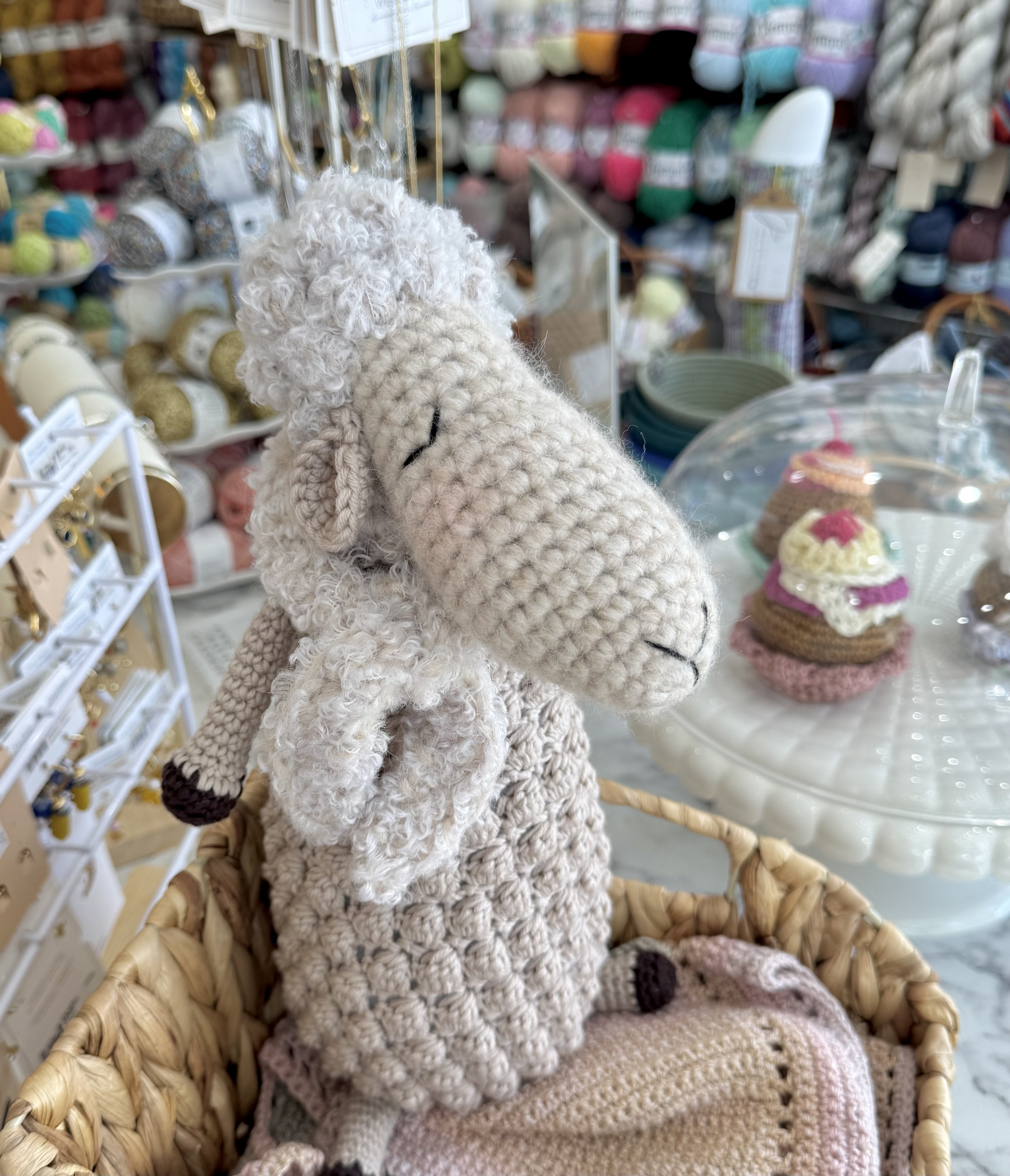 A light beige crocheted sheep doll with curly details, sitting in a wicker basket in a yarn store.
