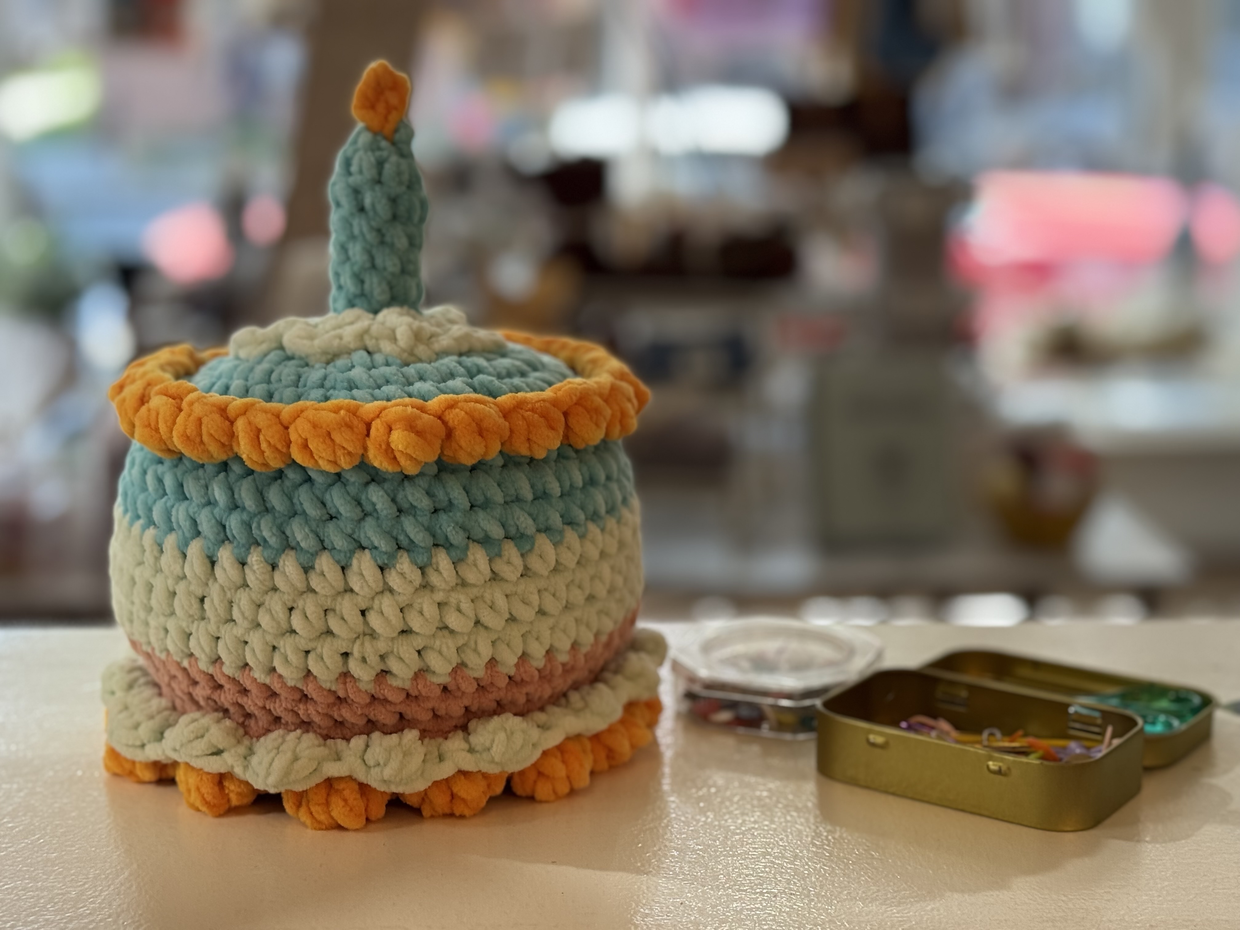 A cake made of blue, pink, white, and orange crochet with an orange candle, placed on a white surface next to small metal boxes.